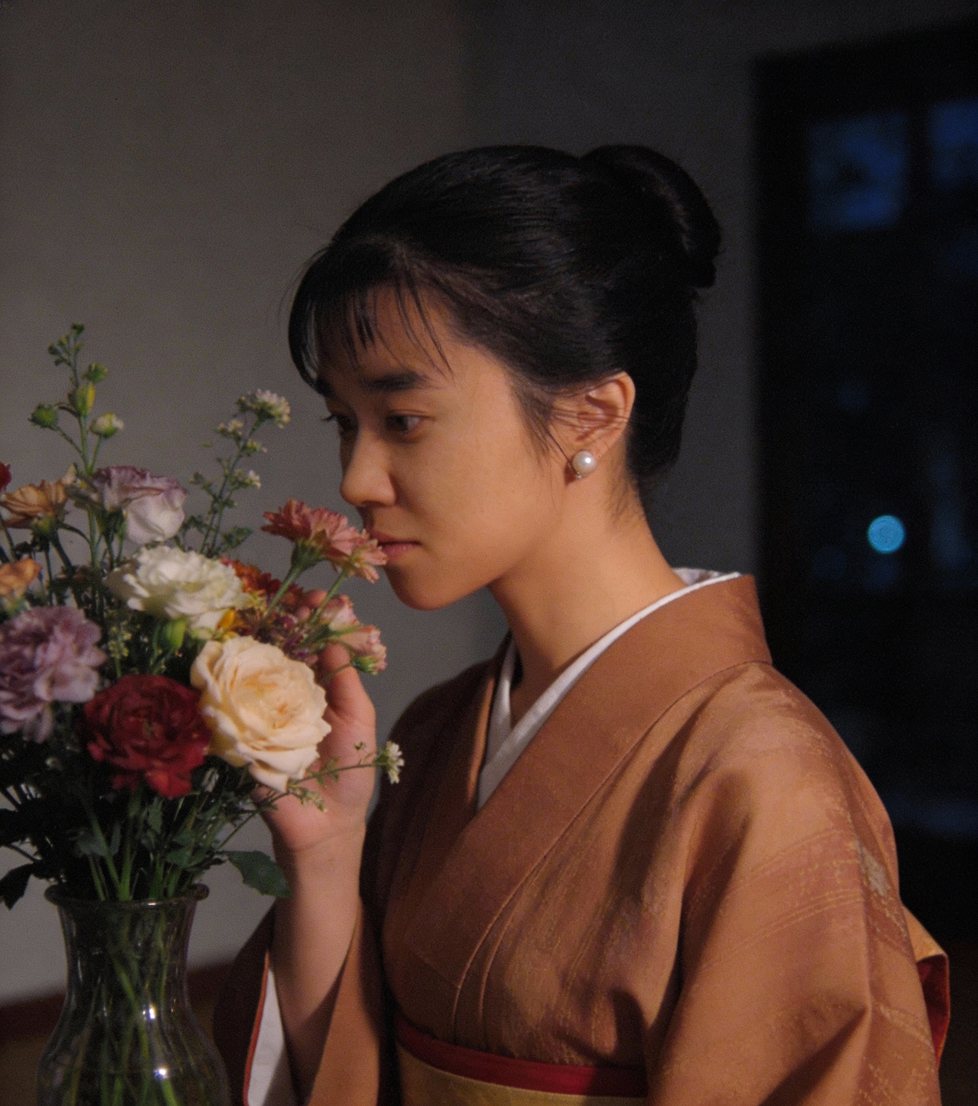 Woman in a peach kimono smelling a bouquet of flowers in a glass vase.