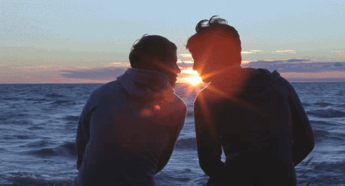 Two people sitting on a beach at sunset, silhouetted with the sun between them over the ocean.